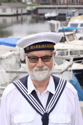 A person with a beard wearing a white sailor hat and a traditional blue and white sailor uniform by a marina.