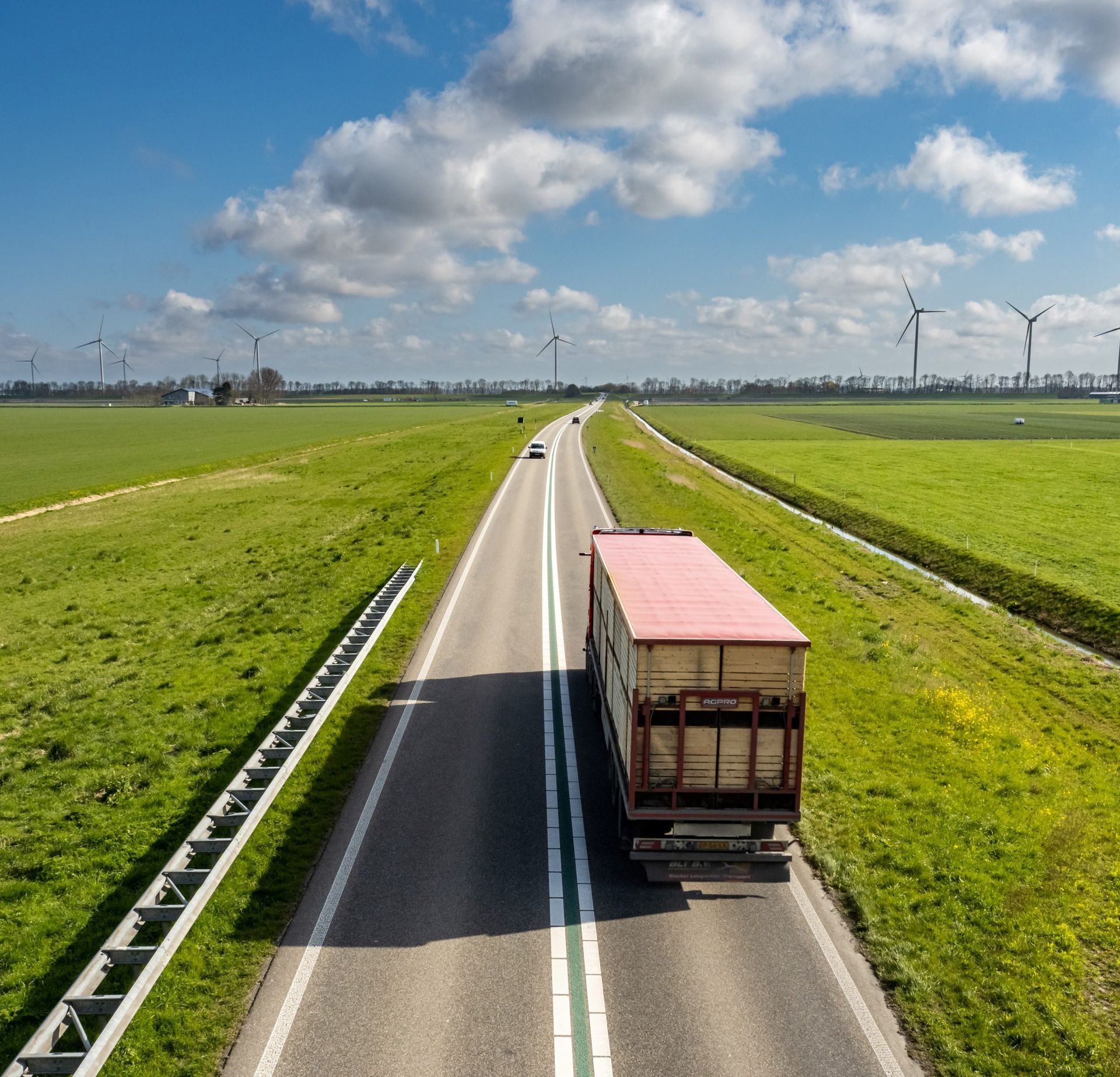 Un camion sur une route, bordée de champs verdoyants, sous un ciel bleu parsemé de nuages. Des éoliennes se dressent au loin.