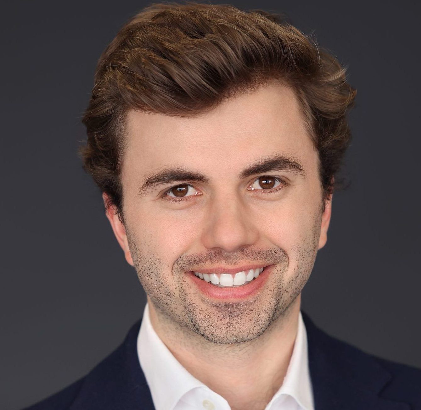 Smiling man in a navy blazer and white shirt against a dark gray background.