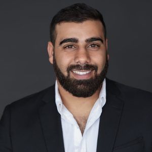 Homme à la barbe foncée, vêtu d'un costume noir et d'une chemise à col blanc, souriant, sur fond gris.