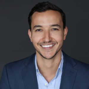 Homme aux cheveux foncés souriant, portant un blazer bleu et une chemise bleu clair sur fond gris.