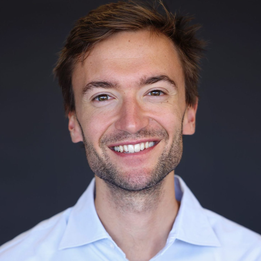 Homme souriant aux cheveux châtain clair, portant une chemise bleue boutonnée, sur fond gris foncé.