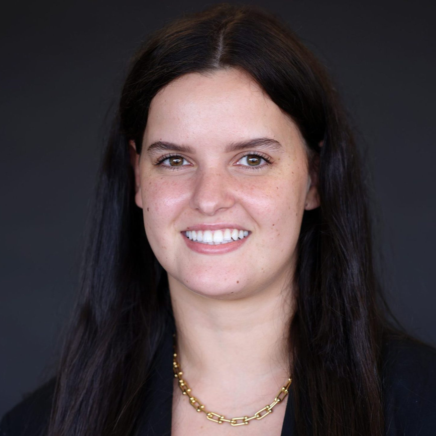 Woman with dark hair, smiling, wearing a gold chain and black blazer, against a dark gray background.