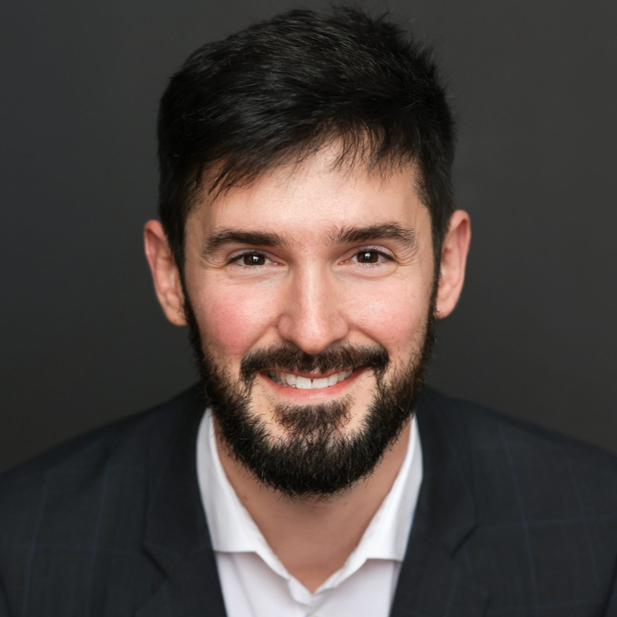 Man in dark suit and white shirt, smiling, against a gray backdrop.