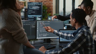 Quatre personnes dans un bureau regardent des écrans d'ordinateur affichant du code. L'une d'elles désigne l'écran du doigt.