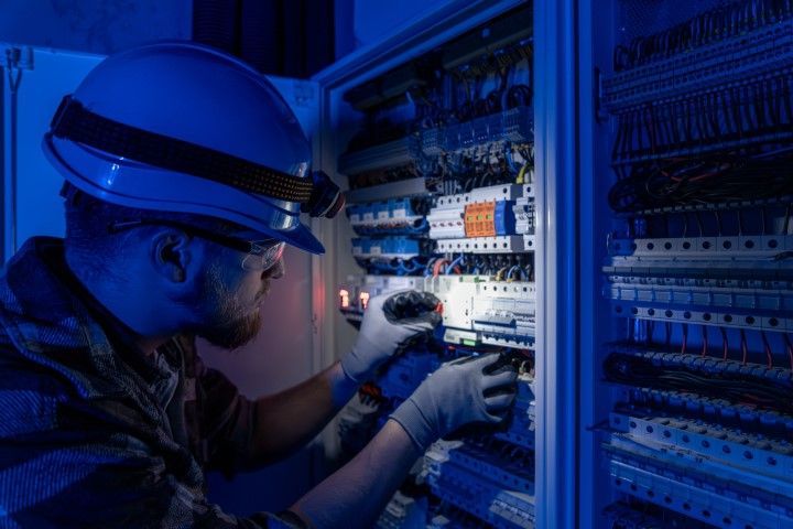 Technician in a hard hat inspecting an electrical control panel lit by blue light