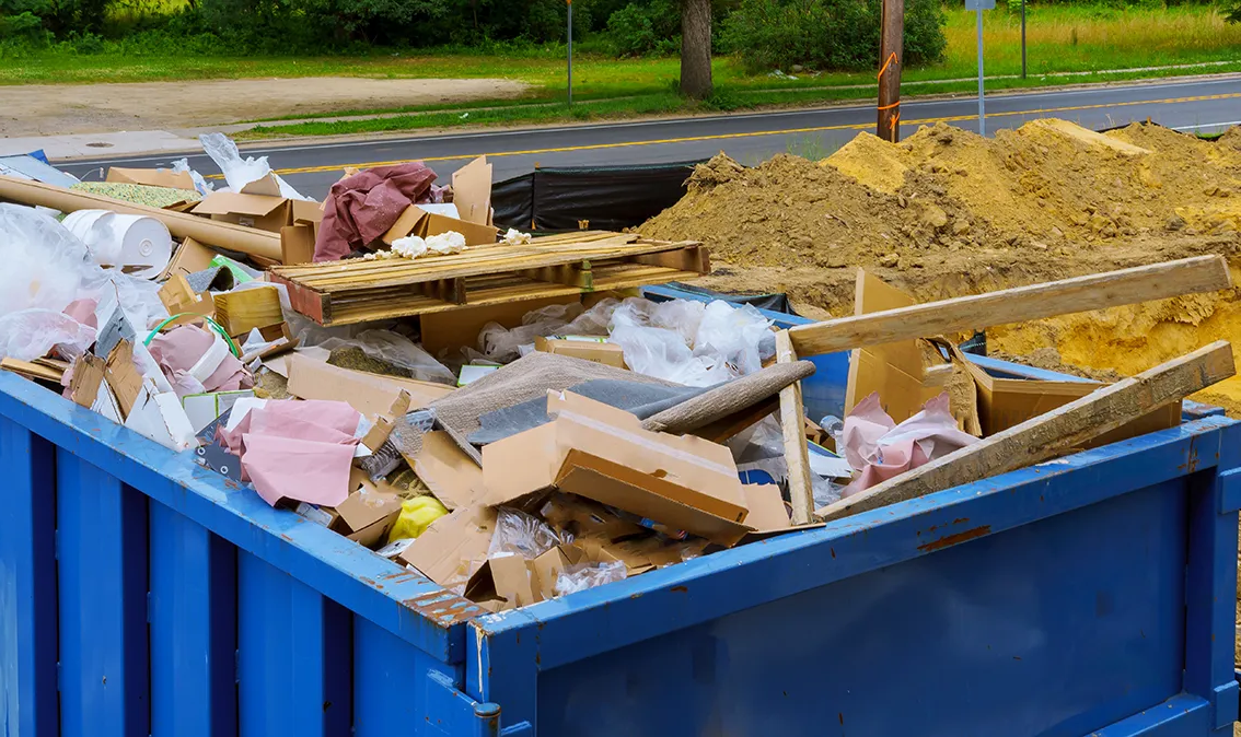 Blue dumpster overflowing with cardboard, wood, and other debris, with dirt pile nearby. — Select Site Solutions in Kiama, NSW