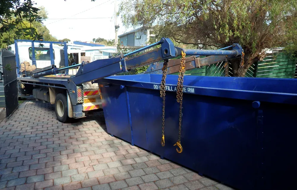 Blue skip bin being lifted by a truck with a crane arm on a brick driveway, outdoor setting. — Select Site Solutions in Kangaroo Valley, NSW