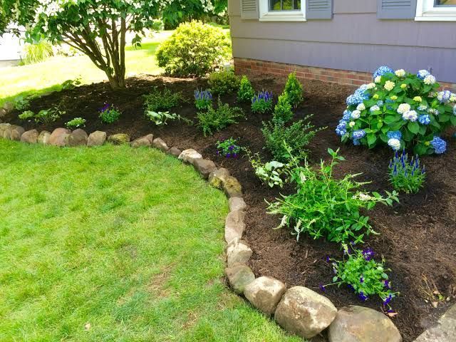 Curved stone border around a mulched flower bed with green shrubs and blue hydrangeas beside a lawn and house.