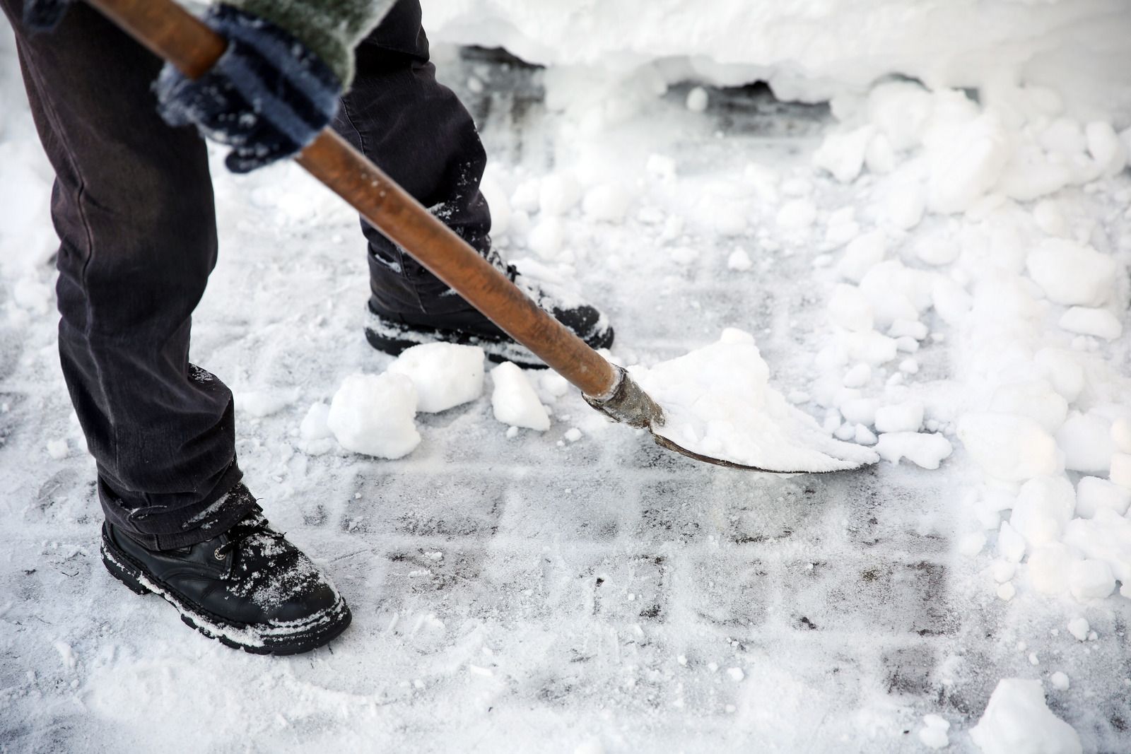 Person shoveling snow from a sidewalk with a wooden shovel handle and black boots