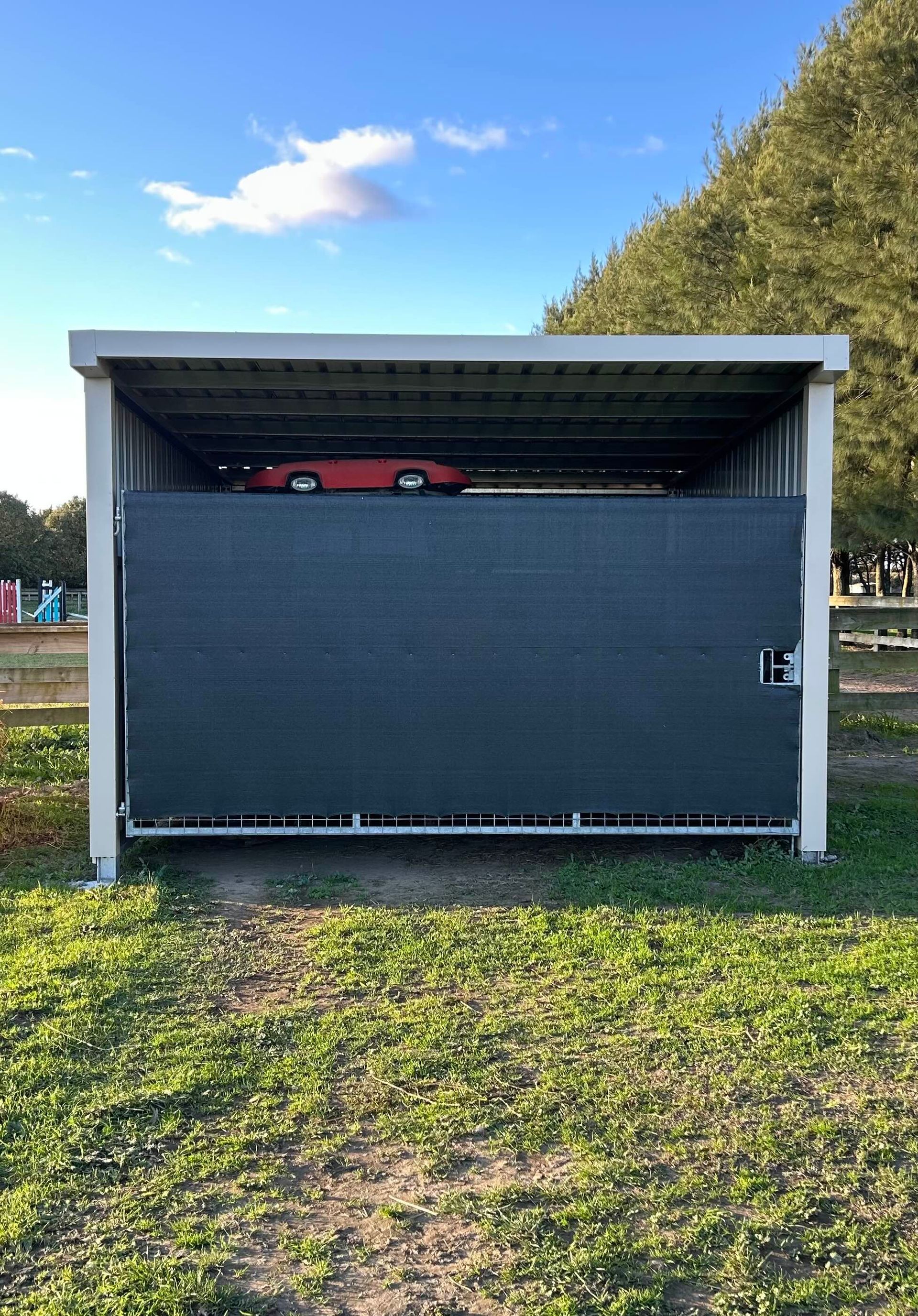 Red car parked under a metal carport with a closed gray door, on a grassy field.