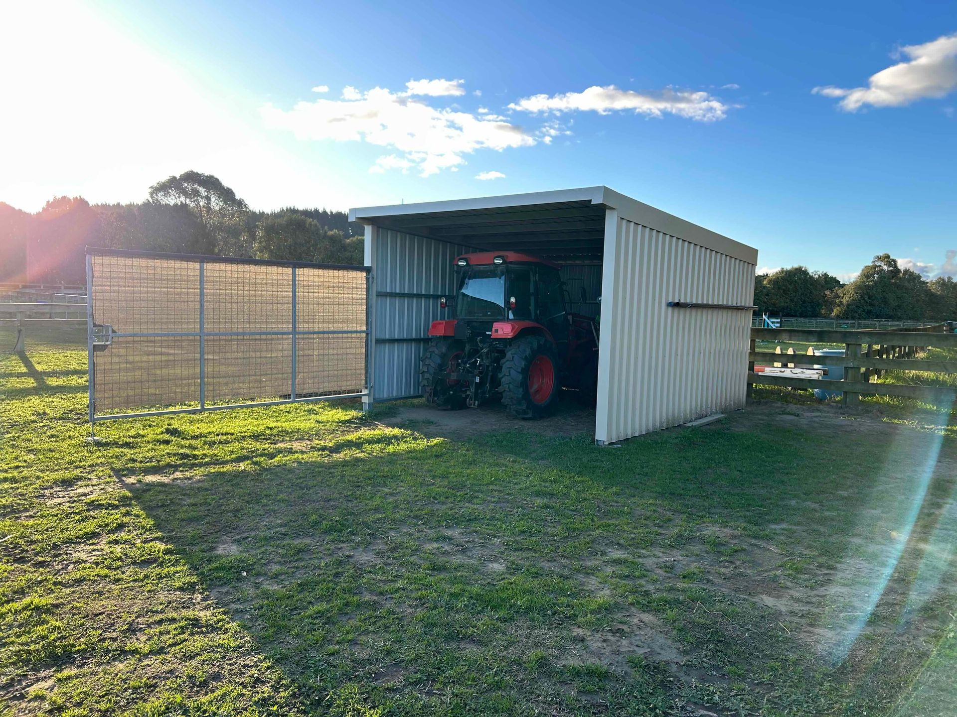 Red tractor parked inside a corrugated metal shed on a grassy field under a blue sky.