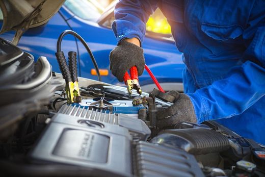 Mechanic using jumper cables on a car battery in a blue engine bay