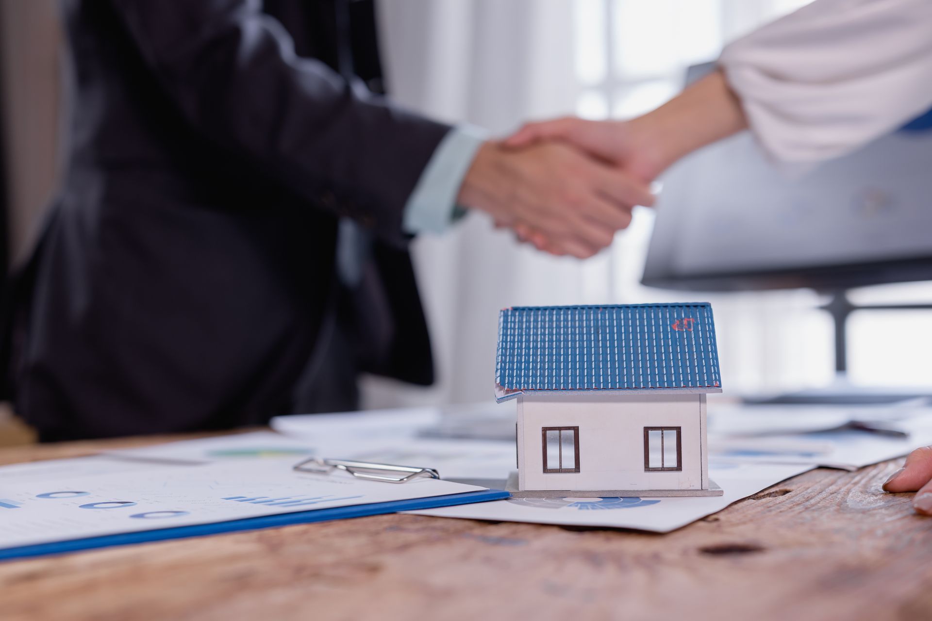 Hands shaking over a house model on blueprints, with a laptop in the background.