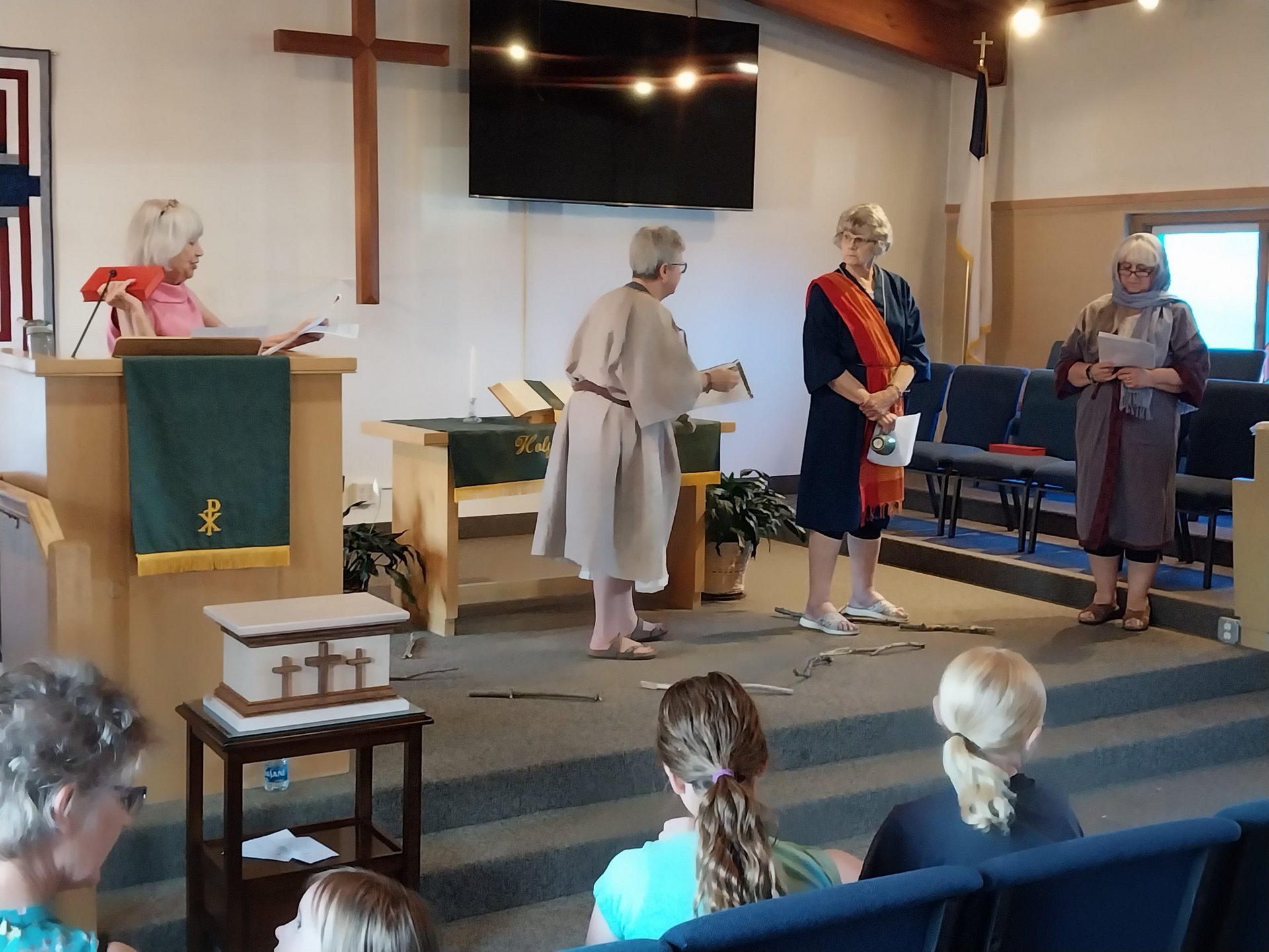 People in a church, performing a play. One woman in robes holding something, other women watching.