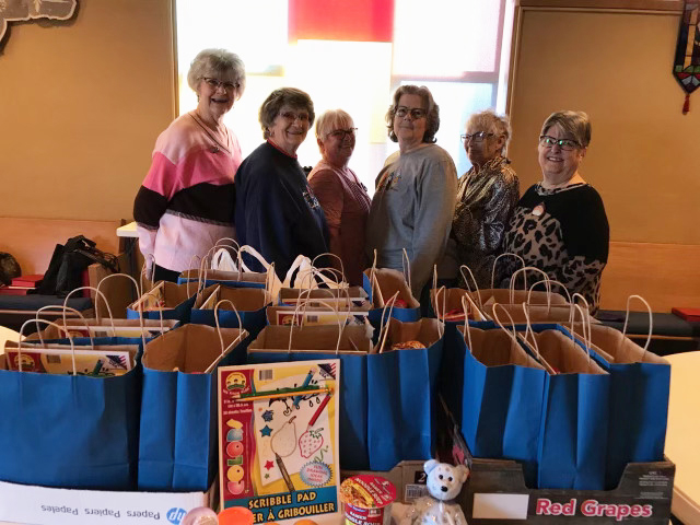 Six women with bags of gifts. Blue bags sit in front of them on a table.