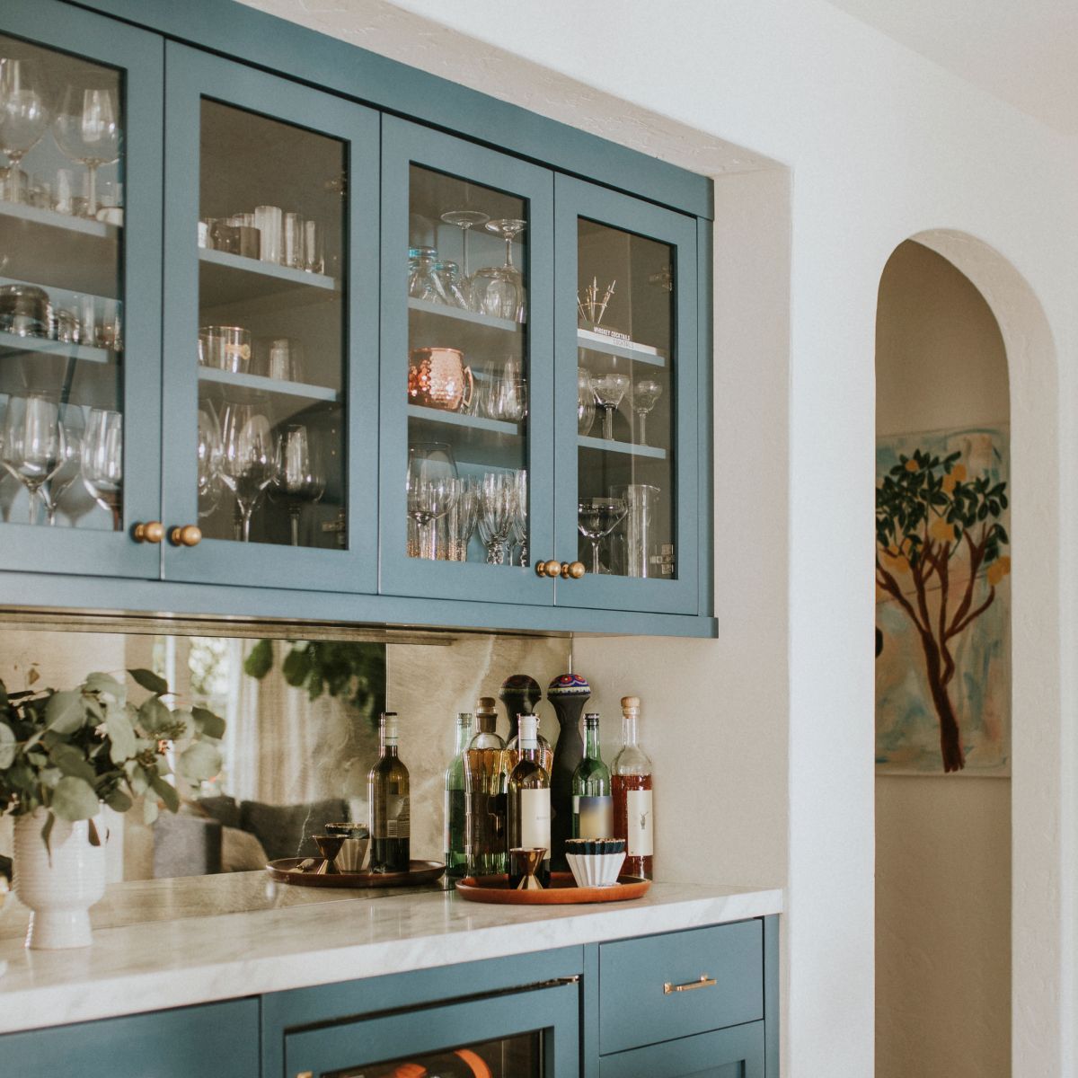 Blue kitchen cabinets with glass doors, white countertop, and an arched alcove with wall art.