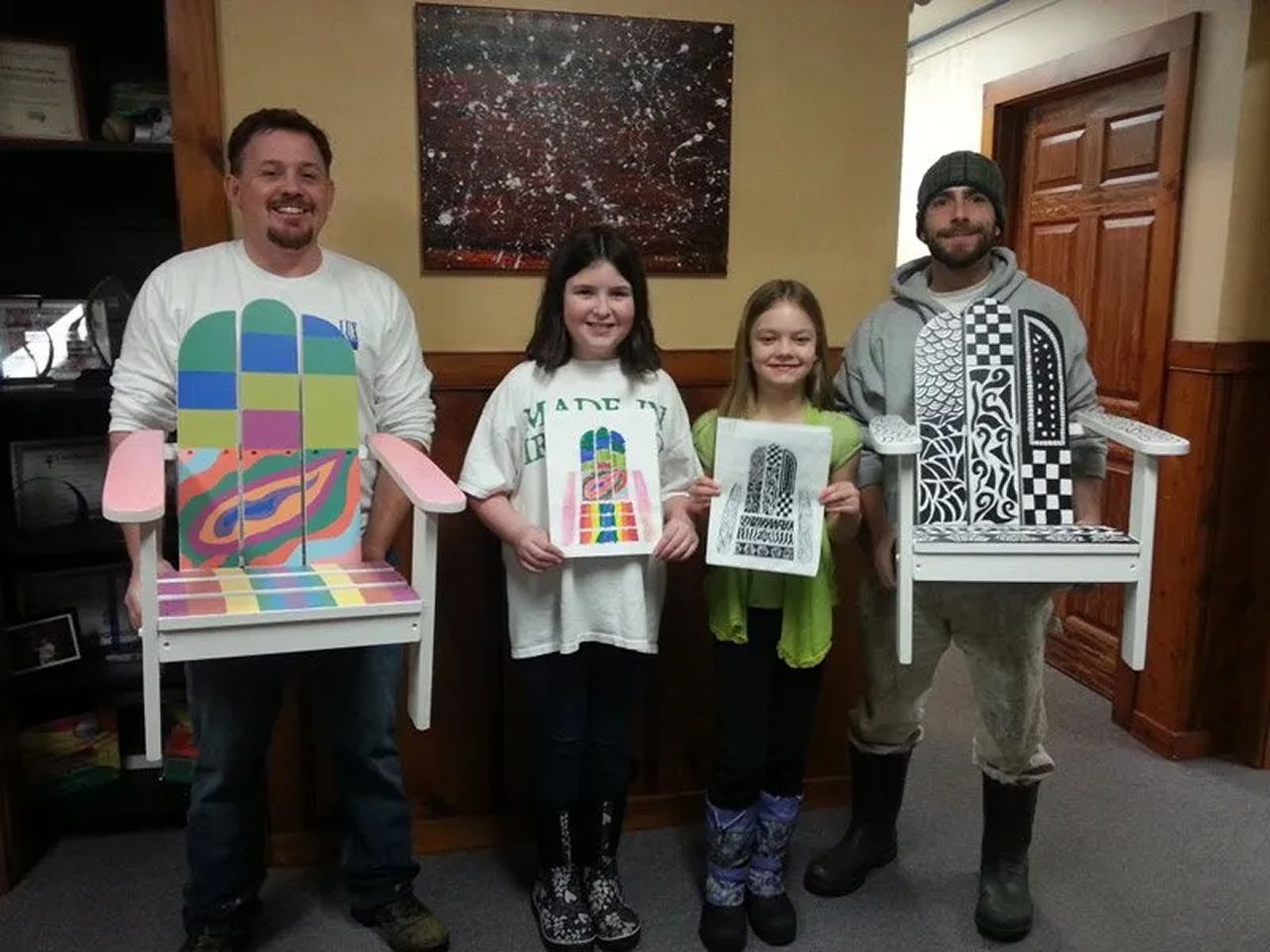 Four people indoors holding colorful art prints and standing in front of a wall painting