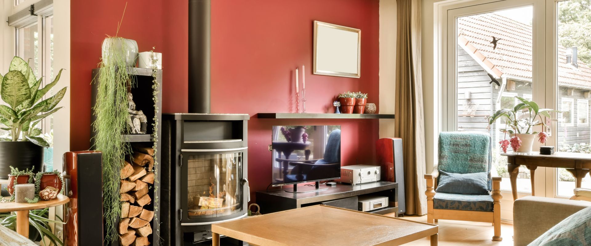Cozy living room with red accent wall, TV, fireplace, plants, and a chair by a bright window.