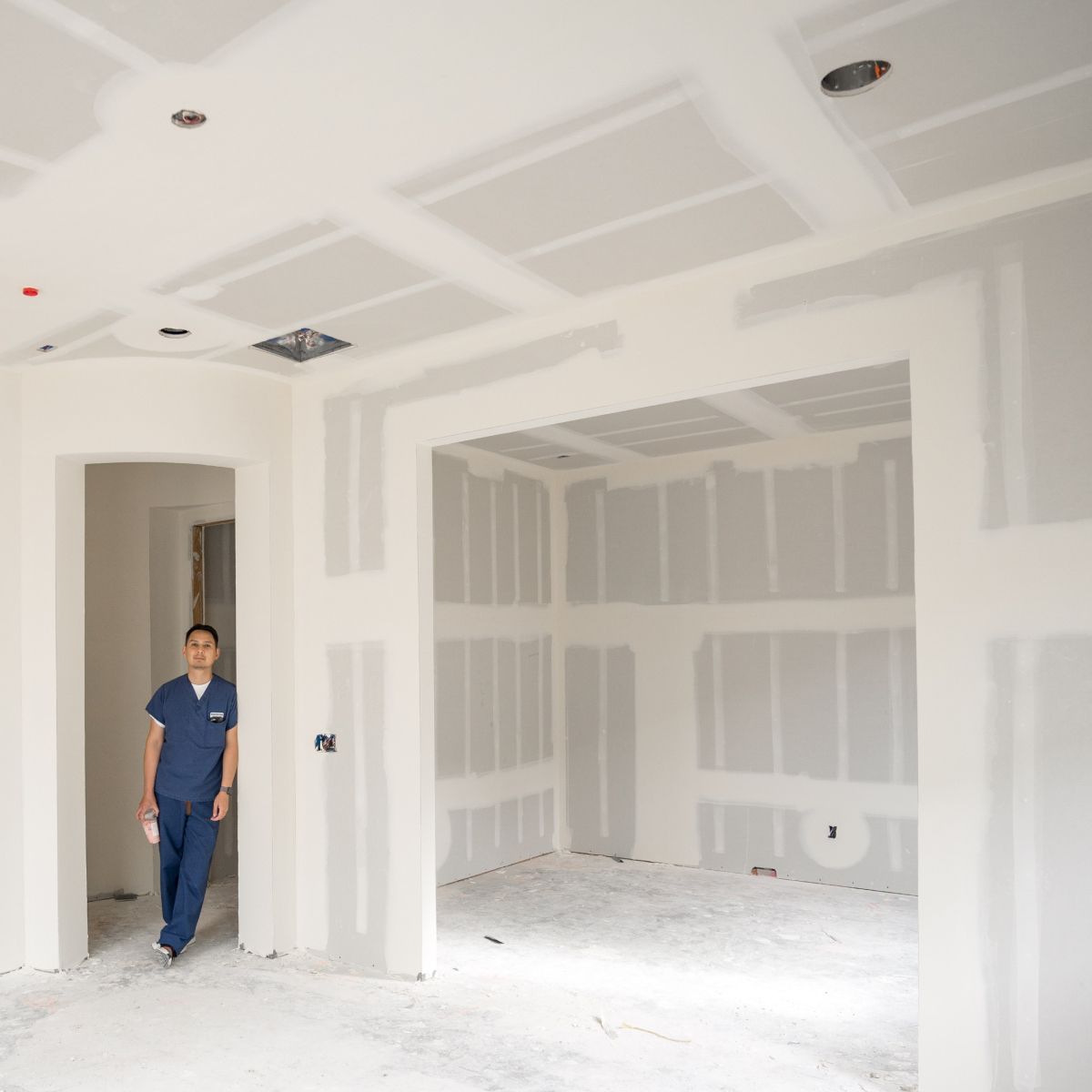 Person in blue coveralls standing in a white, unfinished drywall interior with taped seams and recessed ceiling lights
