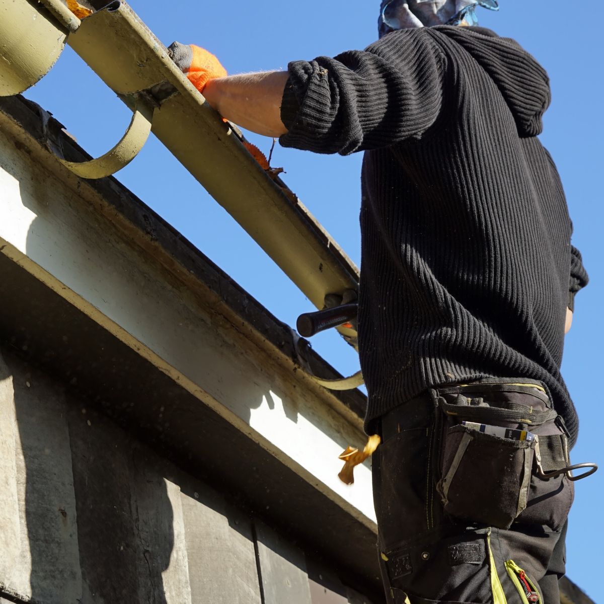 Worker in a black sweater and tool belt painting a building’s roof edge under a blue sky