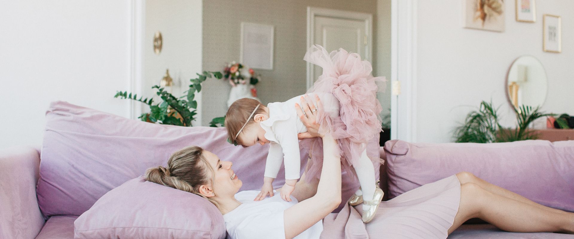 Woman lying on a lavender couch lifts a baby toward her face in a bright living room