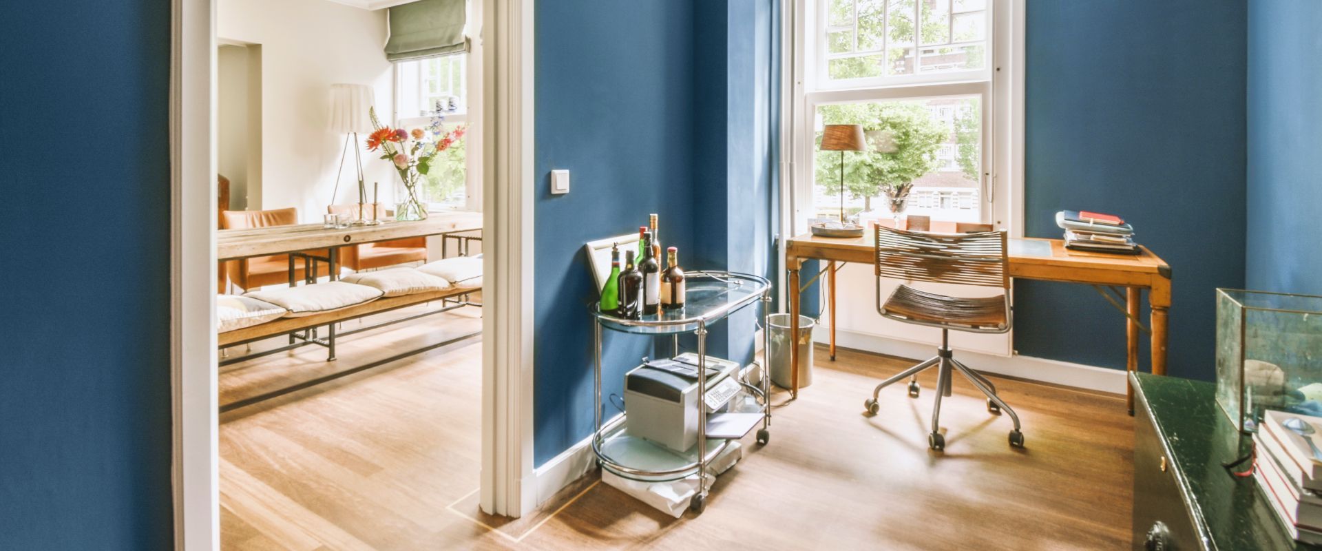 Bright home office with blue walls, wooden desk, chair, shelves, and sunlight through windows