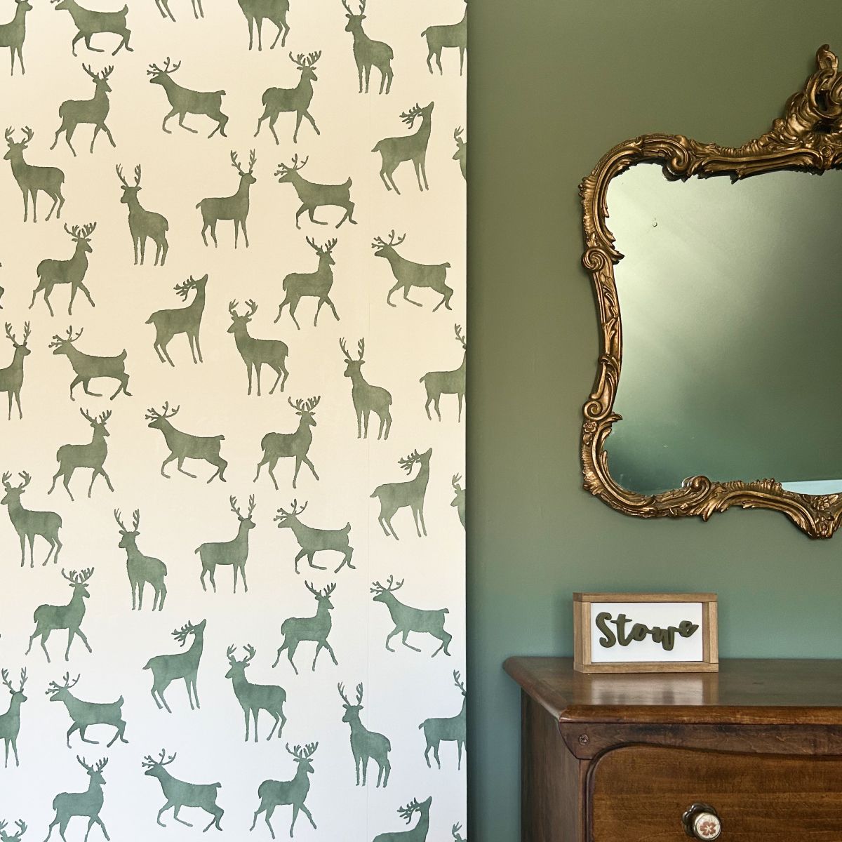 Wall with deer wallpaper beside a green wall, ornate mirror, and small wooden table with framed sign