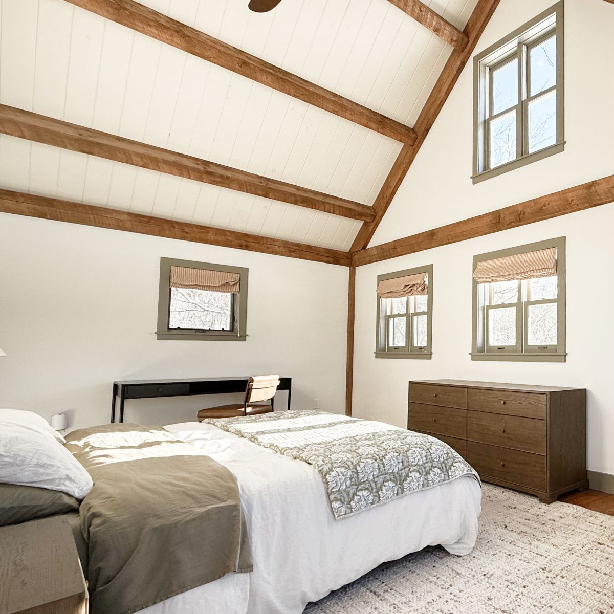 Bright bedroom with vaulted wood-beam ceiling, white bedding, desk, dresser, and two windows