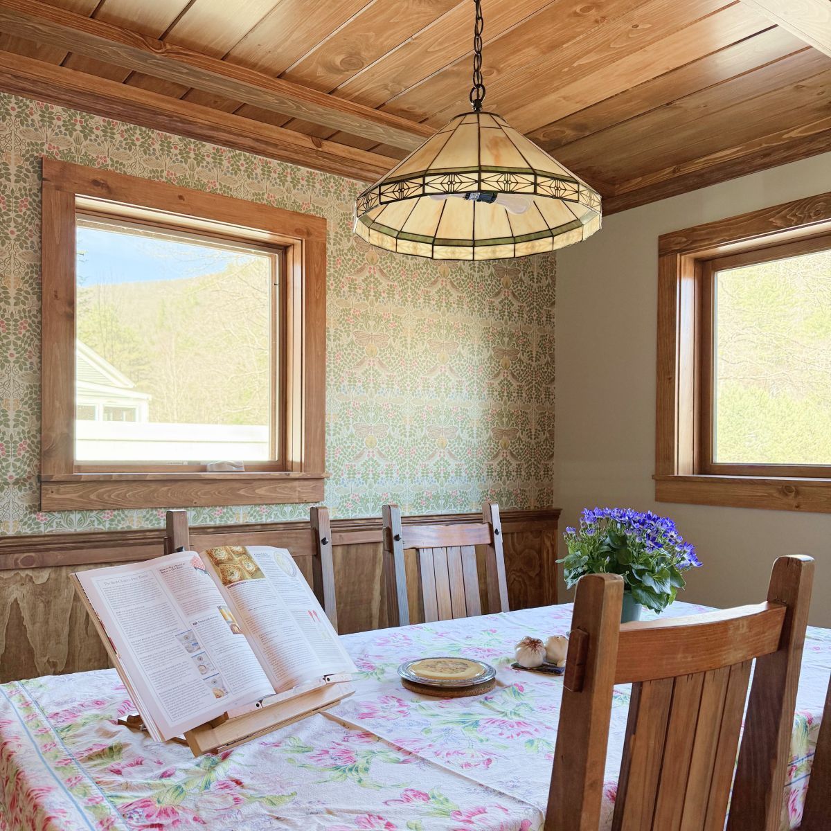 Rustic dining room with floral tablecloth, wooden chairs, stained-glass pendant, and two sunlit windows