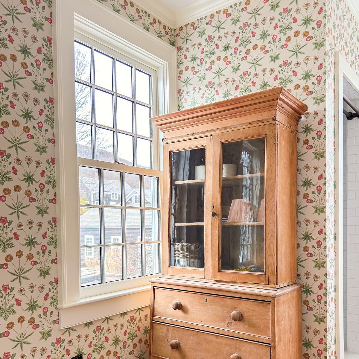 Floral wallpapered room with a wooden glass-front cabinet beside a large window