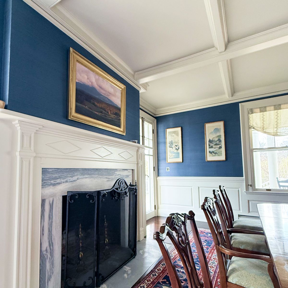 Elegant blue and white dining room with fireplace, framed art, and wooden chairs by a window