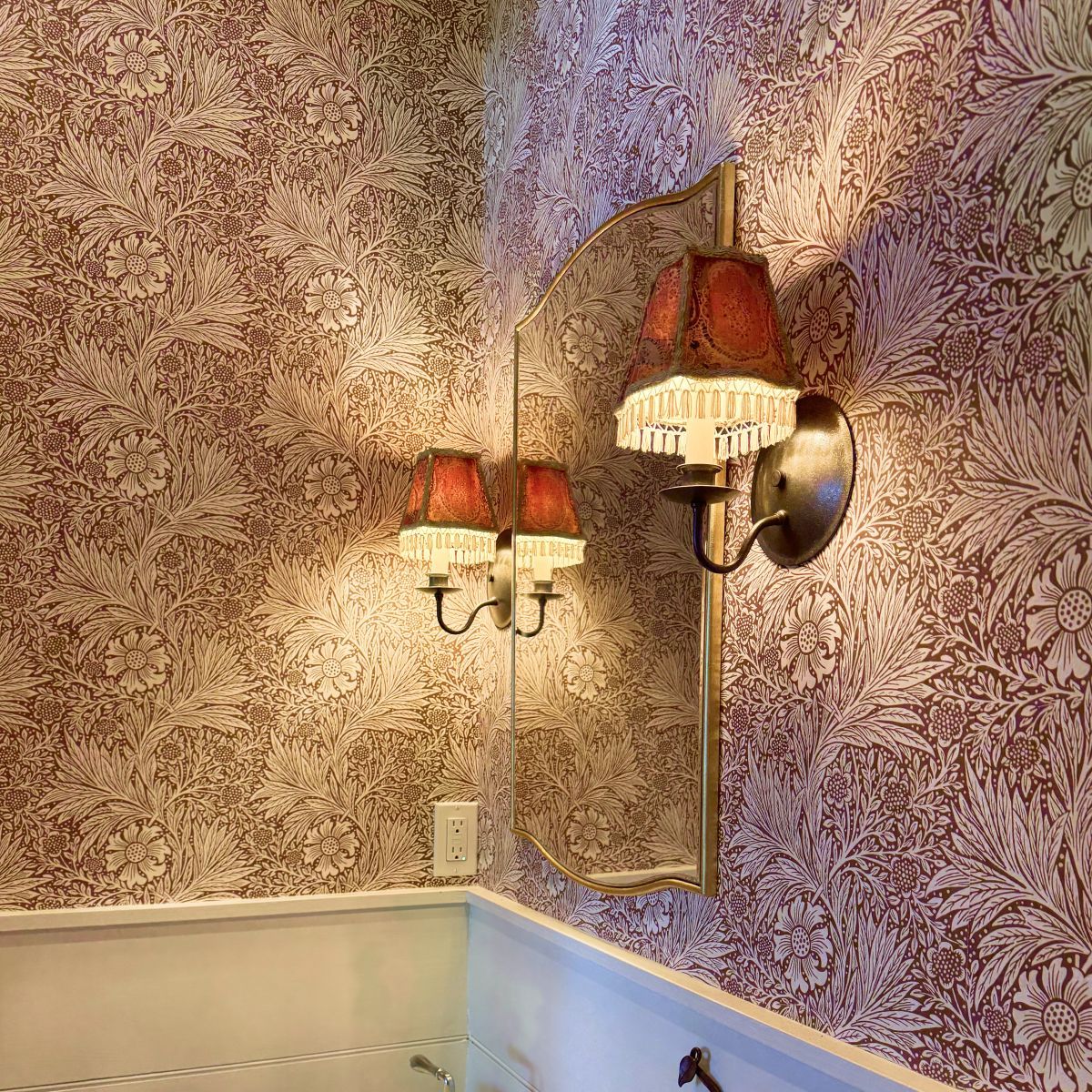 Vintage wall sconces with red shades on patterned wallpaper above white wainscoting