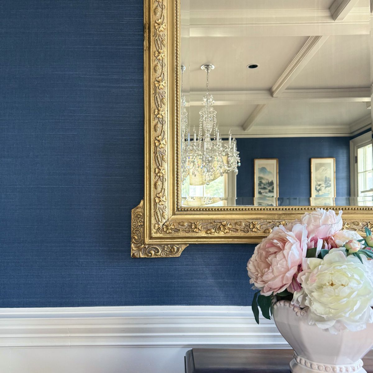 Blue wall with gold-framed mirror reflecting a chandelier, above a white mantel with a pink flower arrangement.