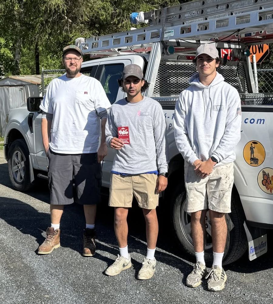 Three men standing beside a white work truck, one holding a red can, in bright sunlight.