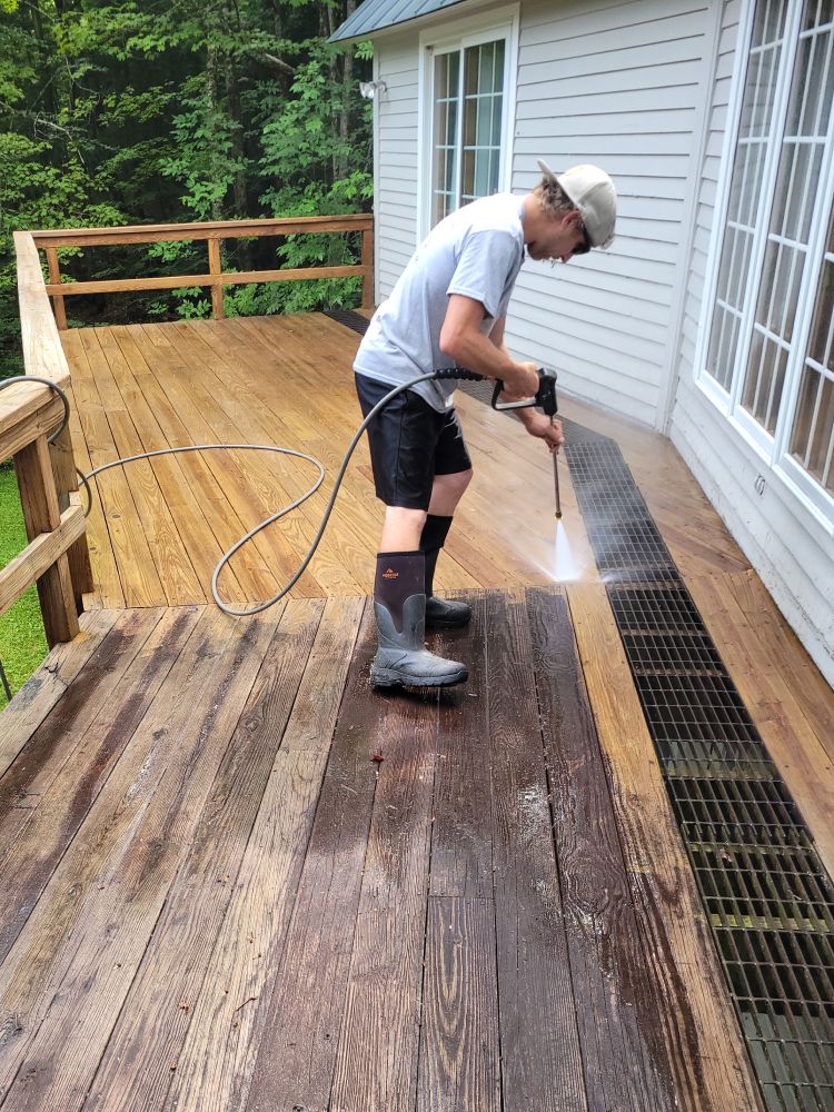 Person pressure-washing a wooden deck beside a house.