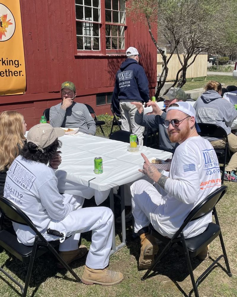 People in white outfits sit at a table outdoors beside a red building, chatting and drinking soda.