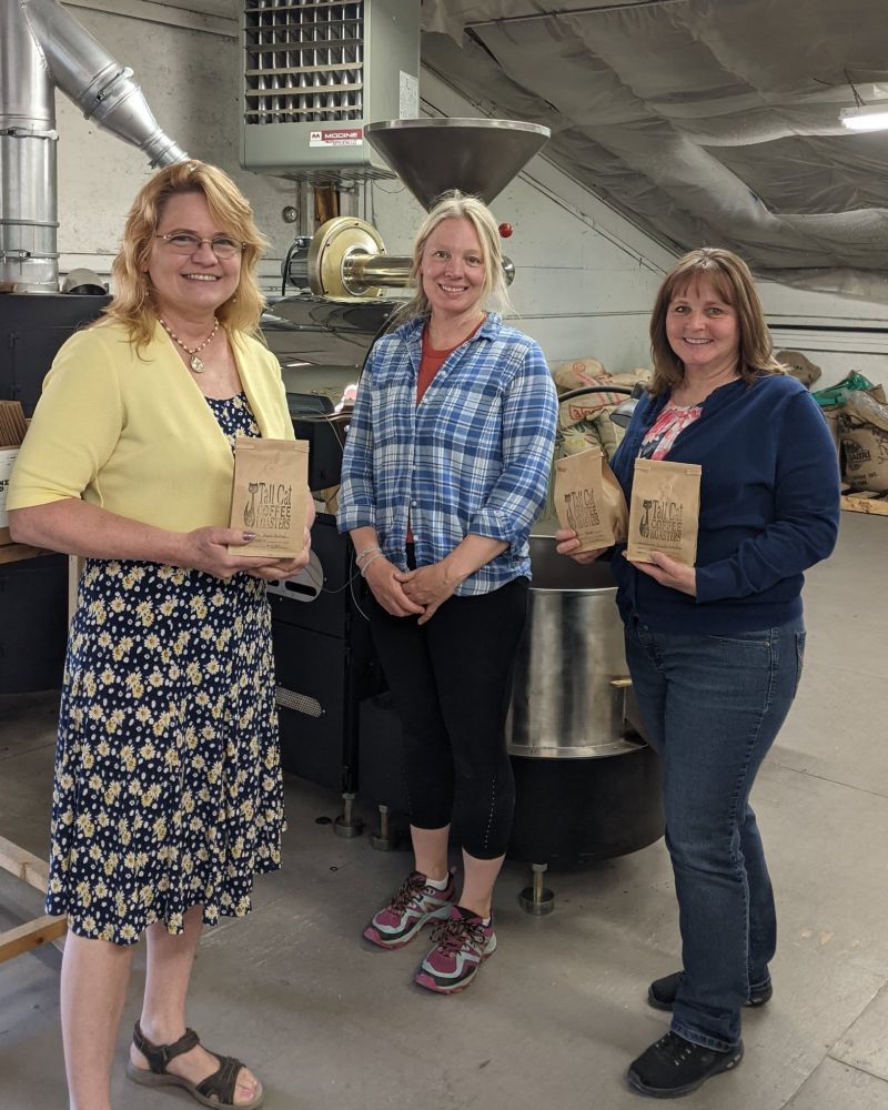 Three people stand in an industrial setting, holding brown paper bags and smiling at the camera.