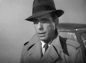 A black and white photo of a man wearing a hat and trench coat.
