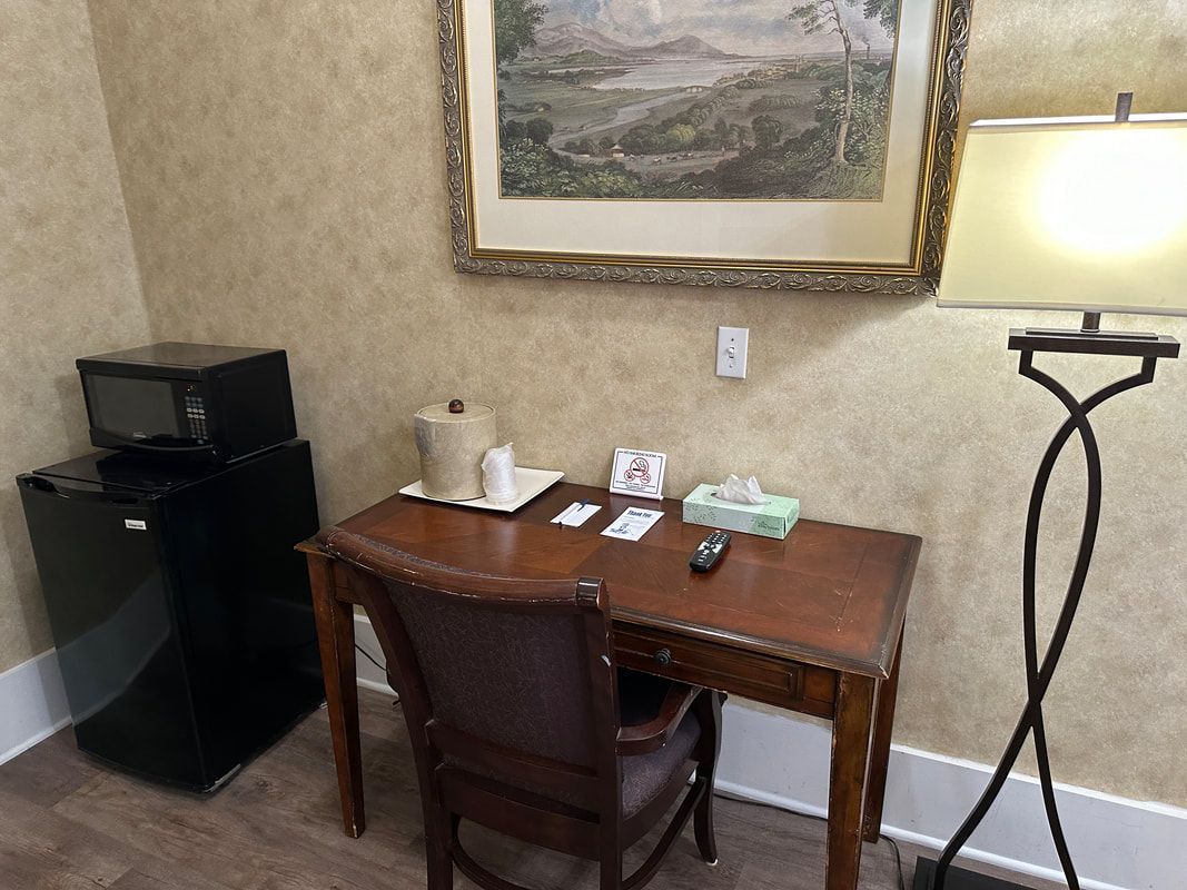 A hotel room with a desk , chair , lamp and microwave.