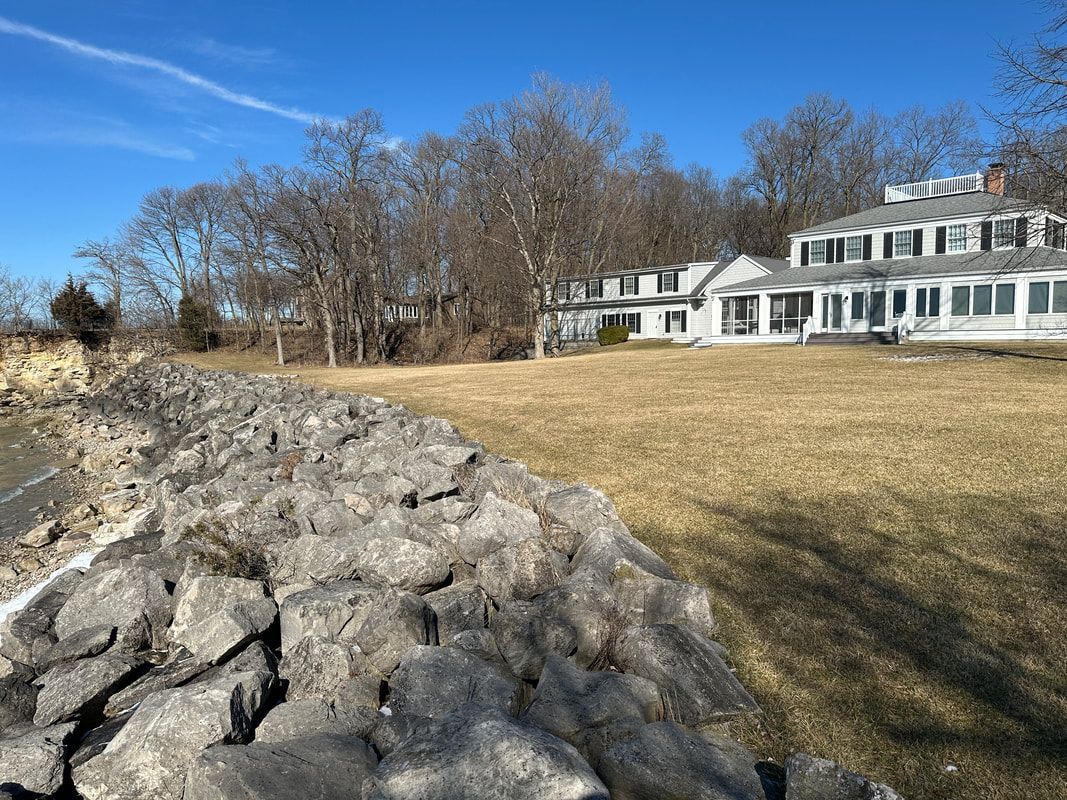 A large white house is sitting on top of a grassy hill next to a body of water.
