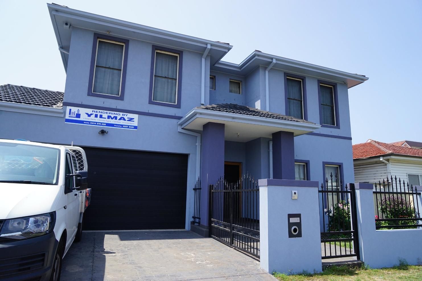 Two-story blue house with a black garage door, gated front yard, and white van parked in the driveway — Rendering By Yilmaz P/L In Wollongong, NSW