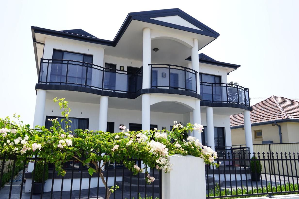 Modern two-story house with white walls, dark blue roof, and black balcony railing behind a fence and flowering hedge — Rendering By Yilmaz P/L In Wollongong, NSW