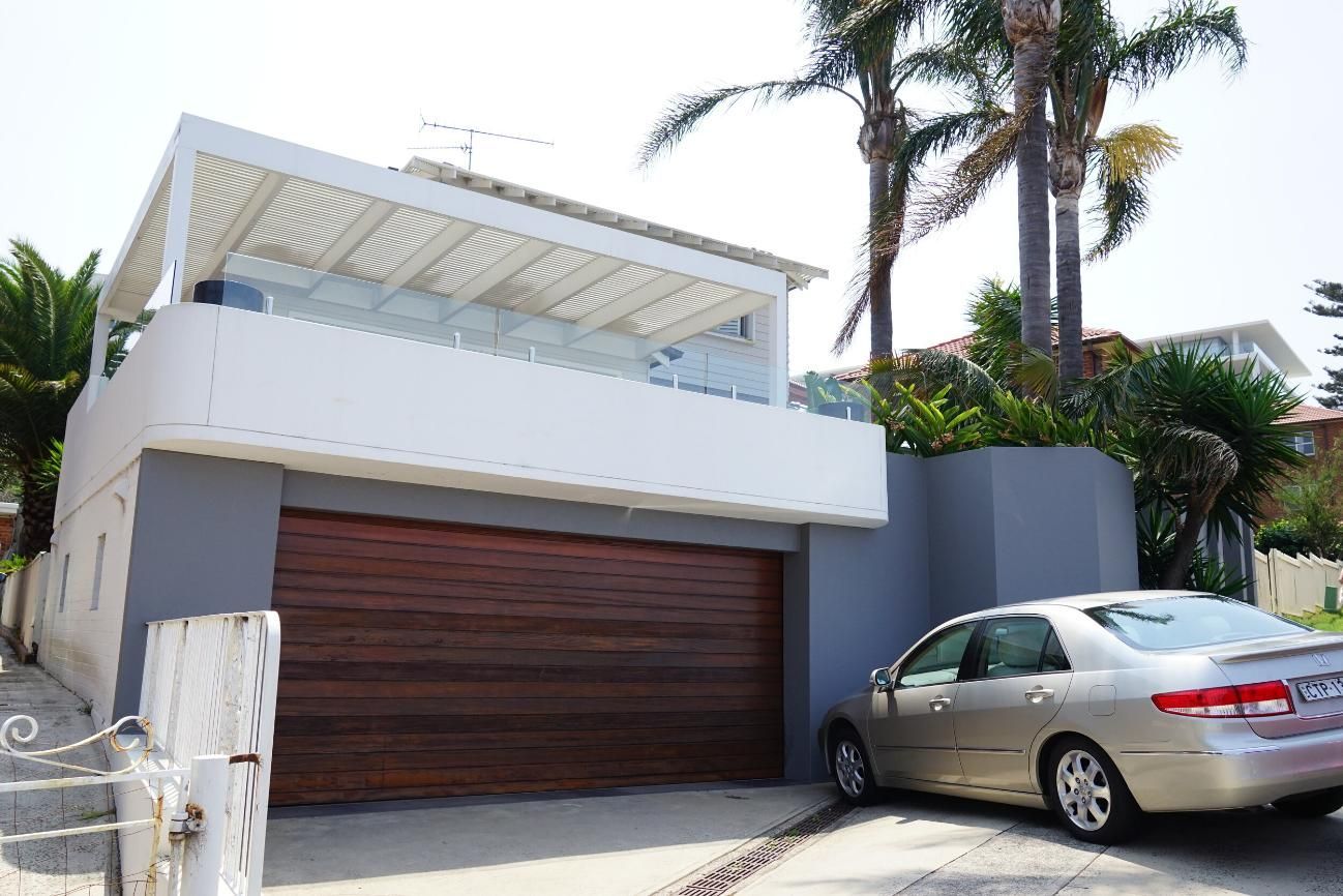 Modern house exterior with a wood garage door, white balcony, palm  trees, and a parked silver car — Rendering By Yilmaz P/L In Kiama, NSW