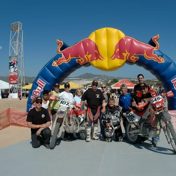 Group of motorcyclists posing under a Red Bull arch at an outdoor event, with bikes in front.