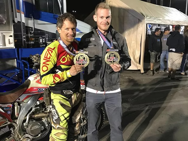 Two men holding trophies beside a motorcycle at a nighttime event
