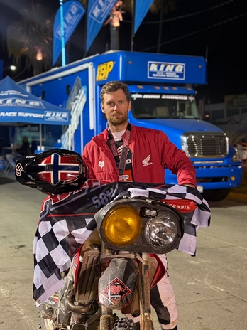 Man in red jacket seated on a scooter, holding a checkered flag in front of a blue truck at night.