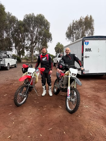 Two dirt bikers pose with red and white motorcycles on a muddy trail near trucks and trees.