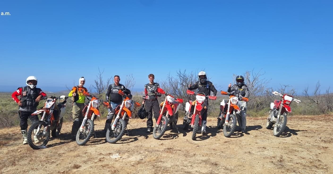 Group of motocross riders posing with dirt bikes on a dry trail under a clear blue sky