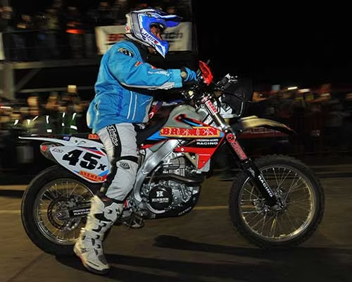 Motocross rider in blue gear on a red dirt bike at night, racing past a crowd